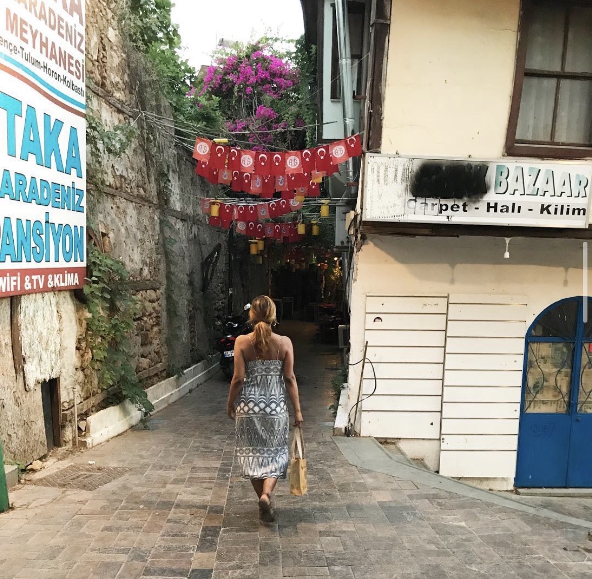 A woman walking down a narrow stone alley in Istanbul, with red Turkish flags and magenta bougainvillea overhead.
